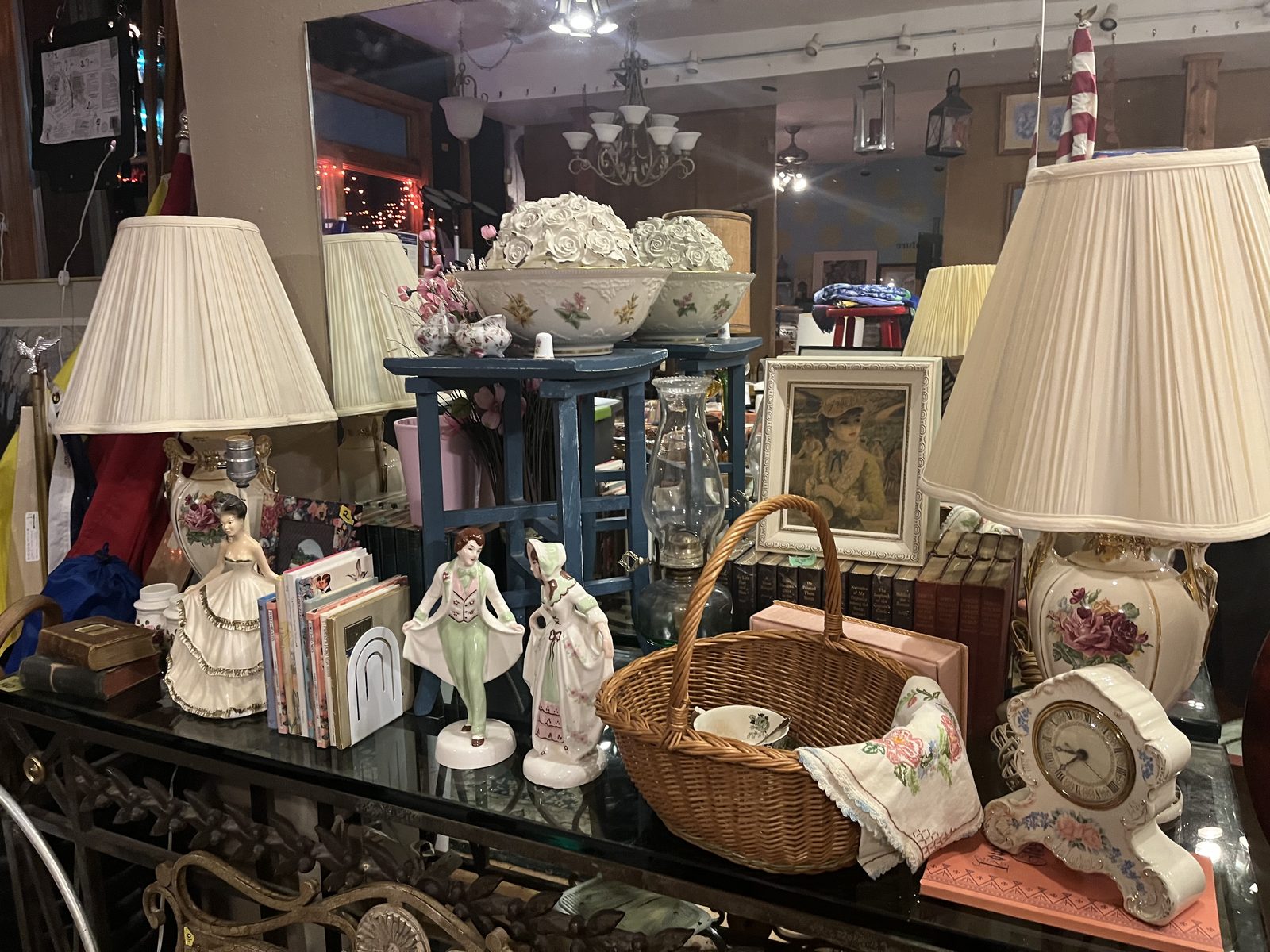 Porcelain figurines, a pair of floral table lamps, a wicker basket, a mantel clock, a framed print and hardcover books on a glass table