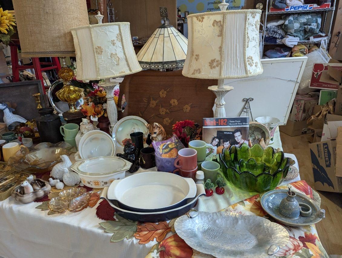 Two table lamps with floral fabric shades on a sale table with green glass, white bakeware, mugs and a stack of CDs