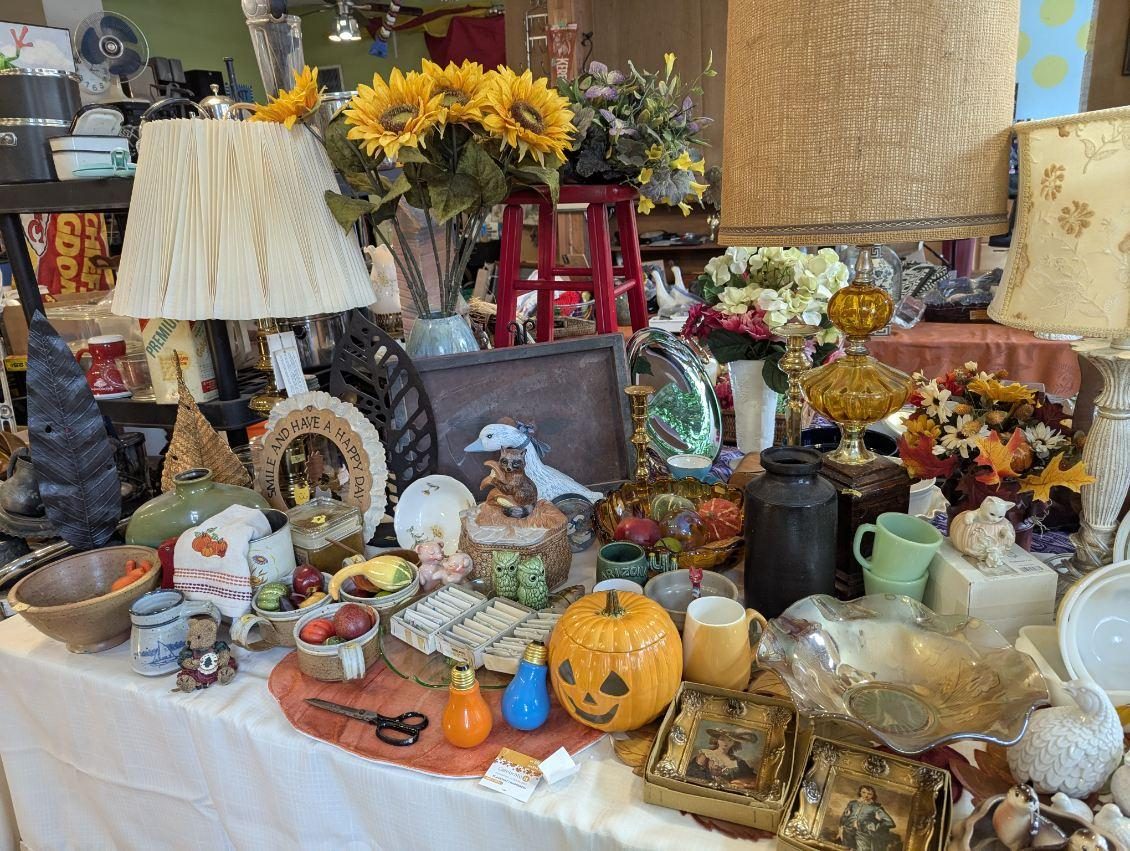 Sale table with a carved jack-o-lantern, sunflower bouquets, a framed picture, brass boxes, stoneware bowls and orange and blue light bulbs
