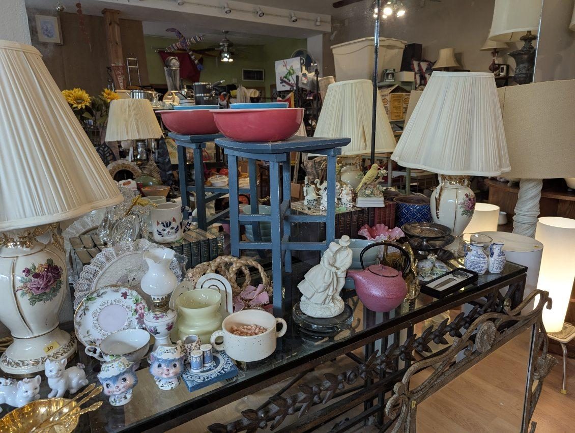 Ornate black metal shelf holding a white floral lamp, china teacups and figurines, a pink teapot and two pink bowls on blue stools