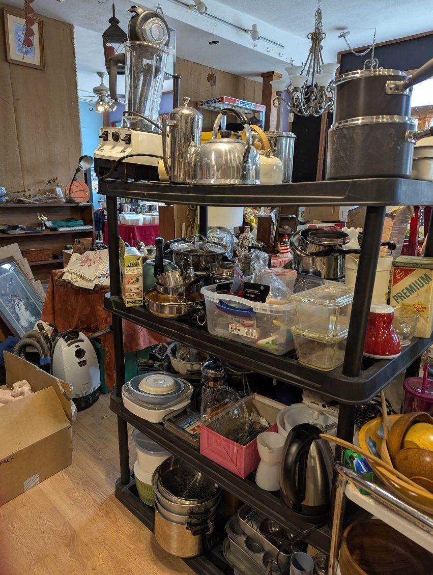 Black shelving unit loaded with stainless pots, a blender, kettles, storage containers and a white carpet cleaner on the floor beside it