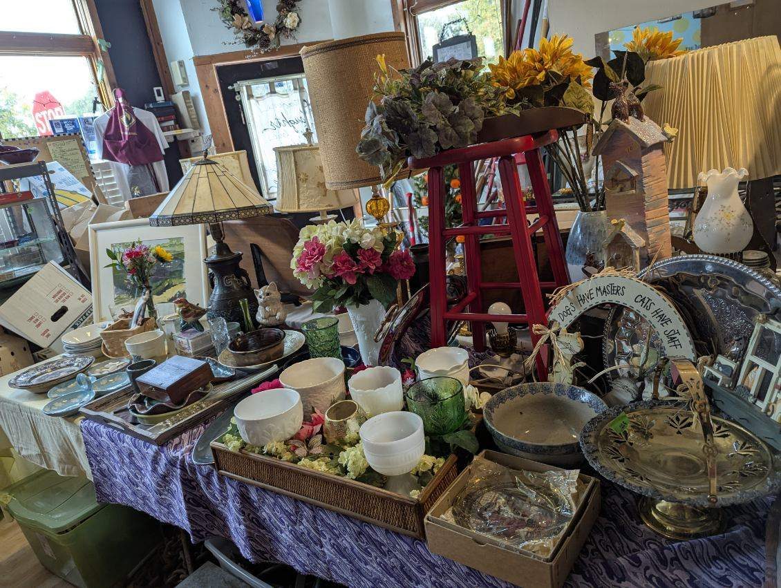 Long sale table by the front window with trays of white pottery, a pressed glass compote, wooden boxes, a red stool and a lamp with a burlap shade